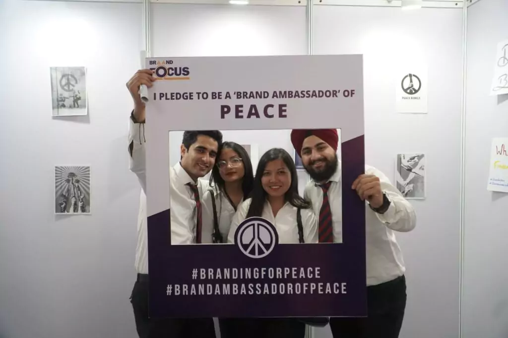 Four standing individuals smiling and holding up a peace sign with their fingers, symbolizing unity and harmony.