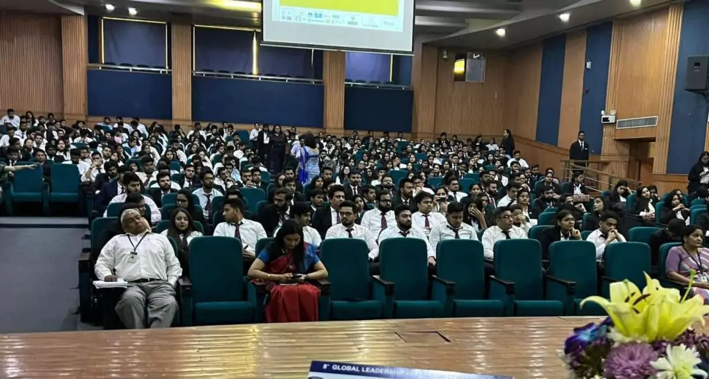 A diverse audience seated in a conference room, listening attentively to a speaker.