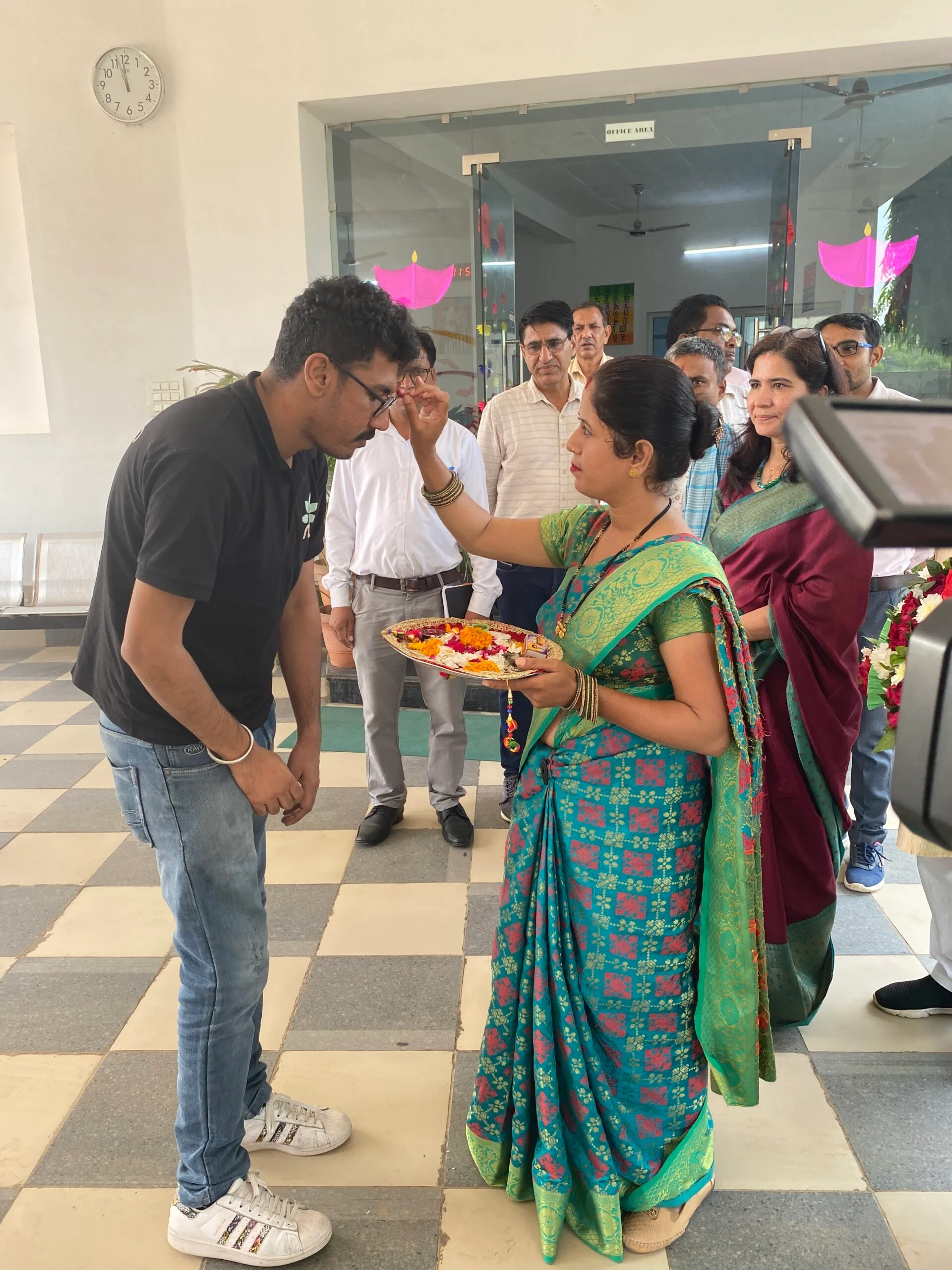 A man and a woman standing in a room, with the lady holding a decorative plate and putting tilak on the man's forehead.