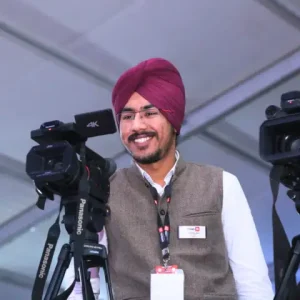 A standing man in a turban, smiling and wearing specs, holding a camera.